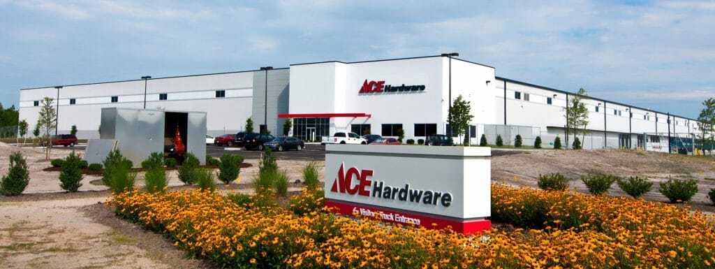 Ace Hardware distribution center with sign and yellow flowers in front, cars parked, and modern building in background.