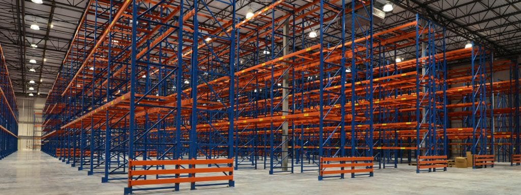 Empty blue and orange warehouse racks, wide view in a spacious industrial storage facility with concrete floor and high ceiling.