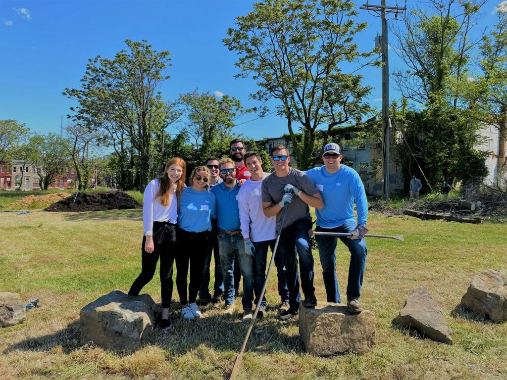 Group of volunteers standing outside on a sunny day, surrounded by greenery and rocks, holding gardening tools, smiling.