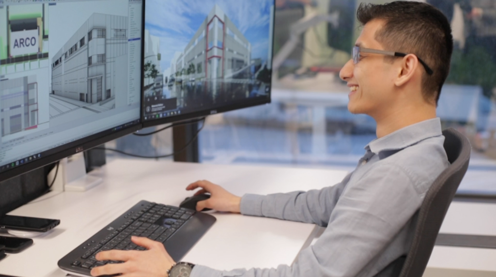 Person smiling at dual monitors with architectural design, wearing a watch, working at a desk, windows in the background.