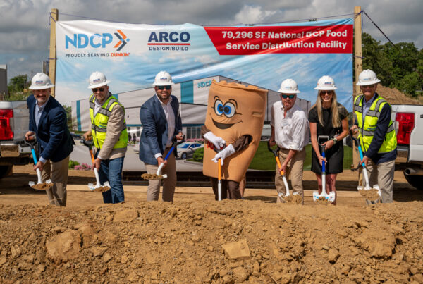 People in hard hats and safety vests at a construction groundbreaking event, with a coffee cup mascot and banner in the background.