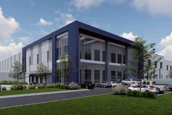 Modern industrial building with blue accents, surrounded by greenery and parked cars under a partly cloudy sky.