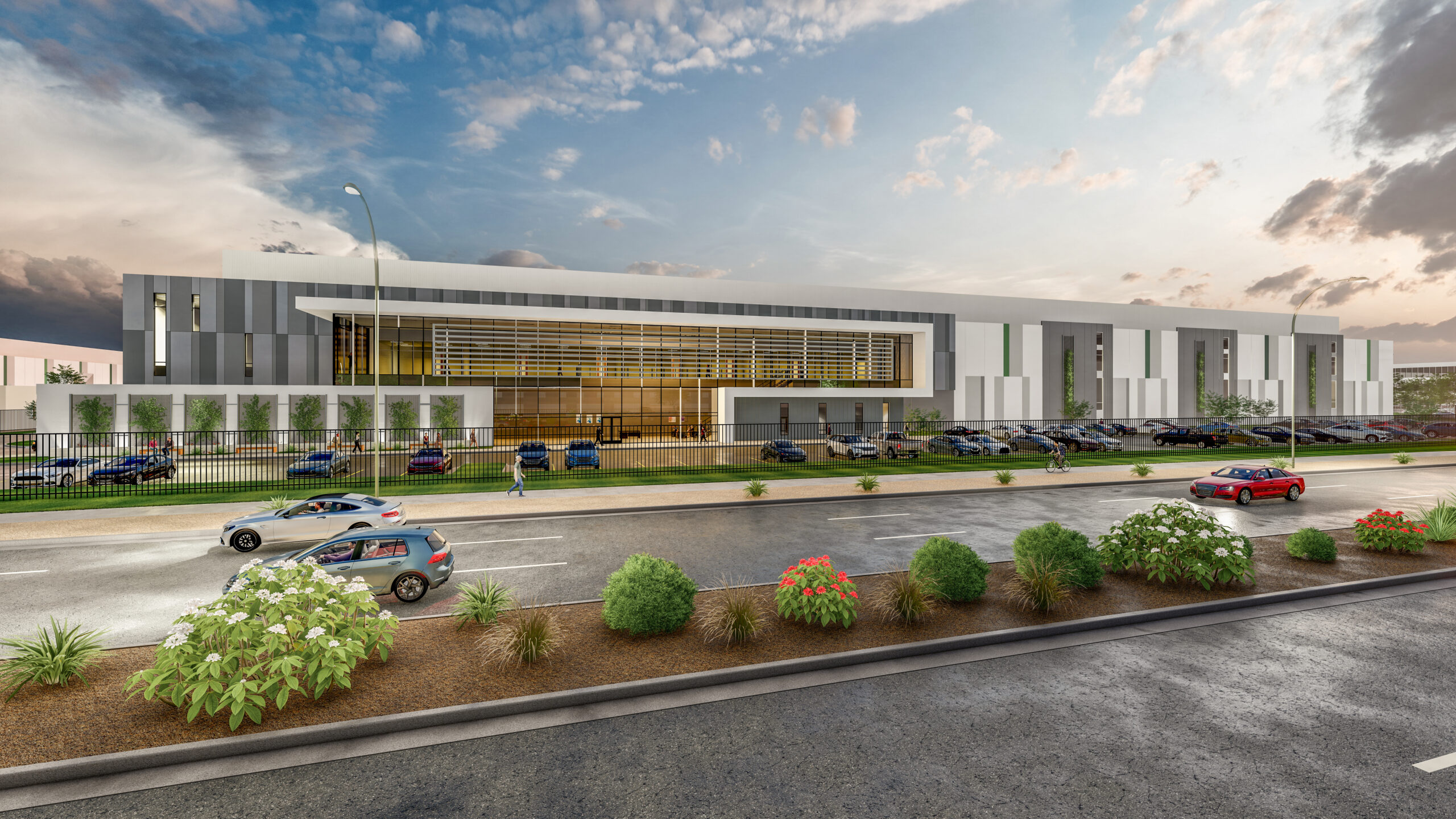 Modern industrial building with parking lot, landscape, and parked cars under a partly cloudy sky at sunset.
